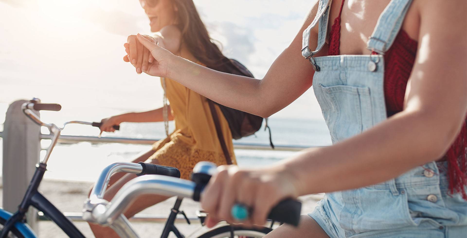 Zwei lächelnde Frauen in entspannter Sommerkleidung genießen eine Fahrradtour entlang der Küste bei sonnigem Wetter.