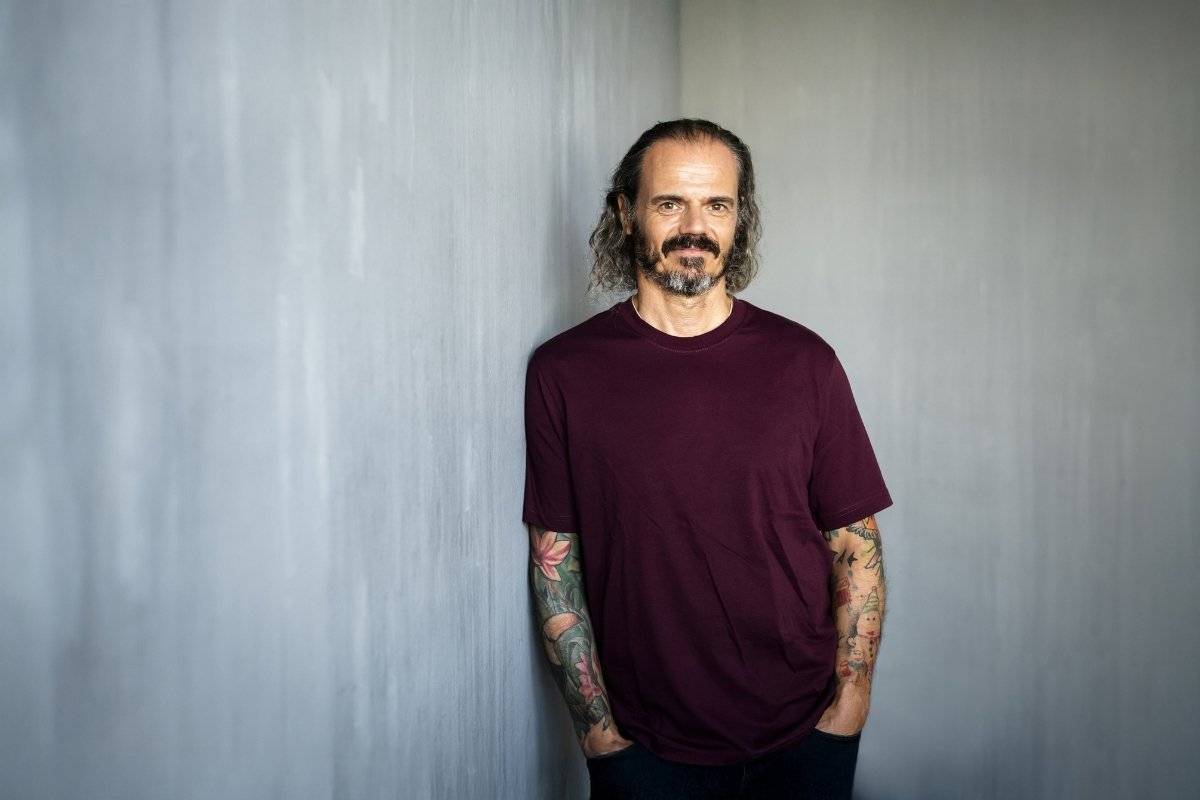 Mann mit schulterlangen Haaren und Bart, trägt ein bordeauxfarbenes T-Shirt, steht entspannt mit verschränkten Armen vor einer grauen Wand, tätowierte Unterarme sichtbar.