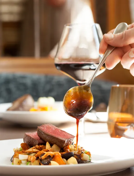 Hand of a chef putting a sauce over a piece of meat on a plate with vegetables, with a glass of red wine on the table.