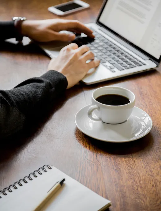 Person working on a laptop with a cup of coffee and a notebook on the table.