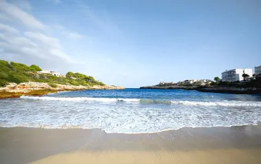Beach with fine sand, clear sea and surrounding houses as well as green nature under a bright blue sky on Mallorca.