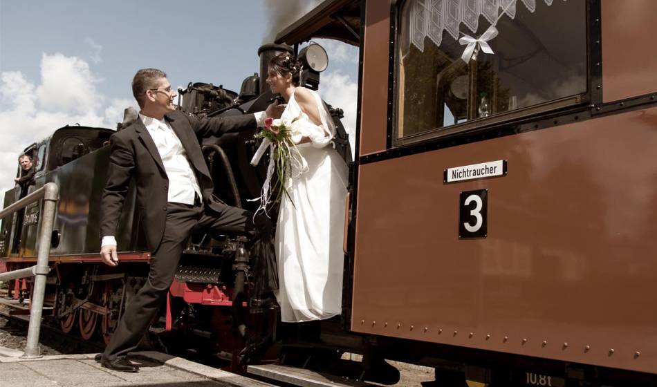 Hochzeitspaar in festlicher Kleidung vor nostalgischer Dampfeisenbahn auf Rügen, einzigartiges Hochzeitsmotiv.