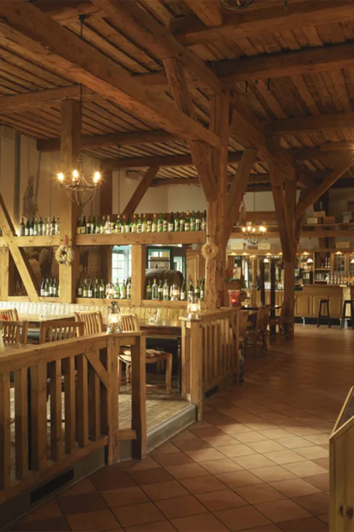 Rustic restaurant with exposed beams and wooden furniture, decorated with wine bottles, lit in warm light.