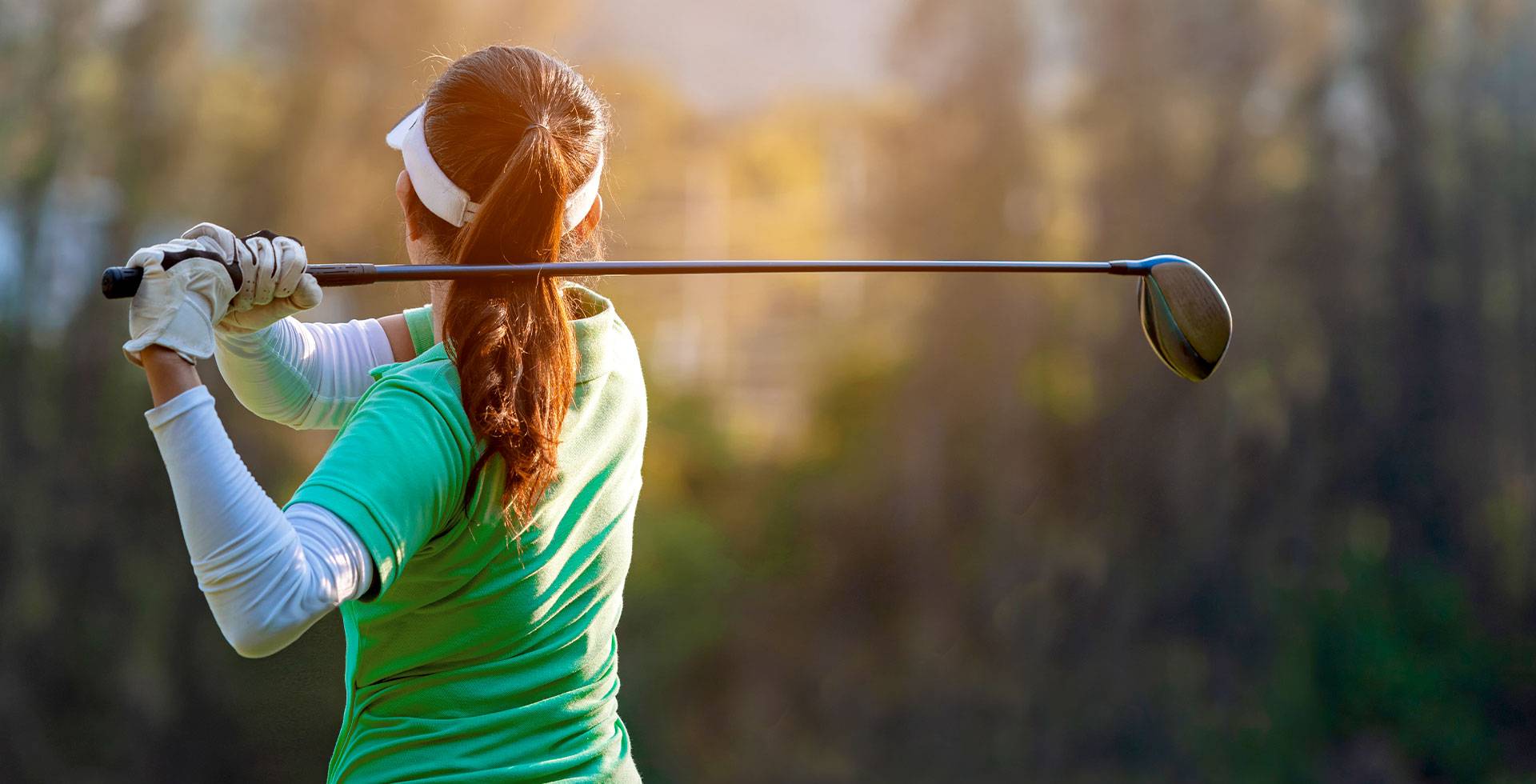 Golferin in grüner Kleidung schlägt mit einem Golfschläger ab, umgeben von Natur und weichem Sonnenlicht.