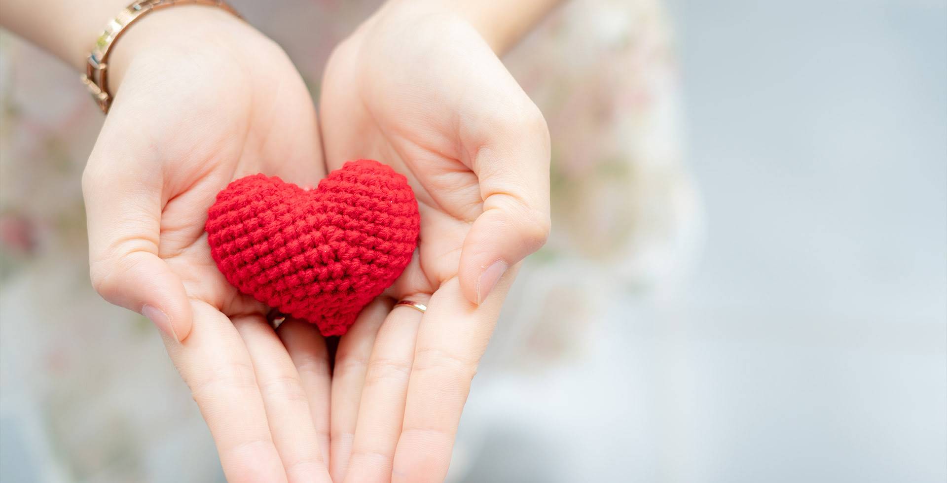 Two hands hold a crocheted red heart as a symbol of love, care and support.
