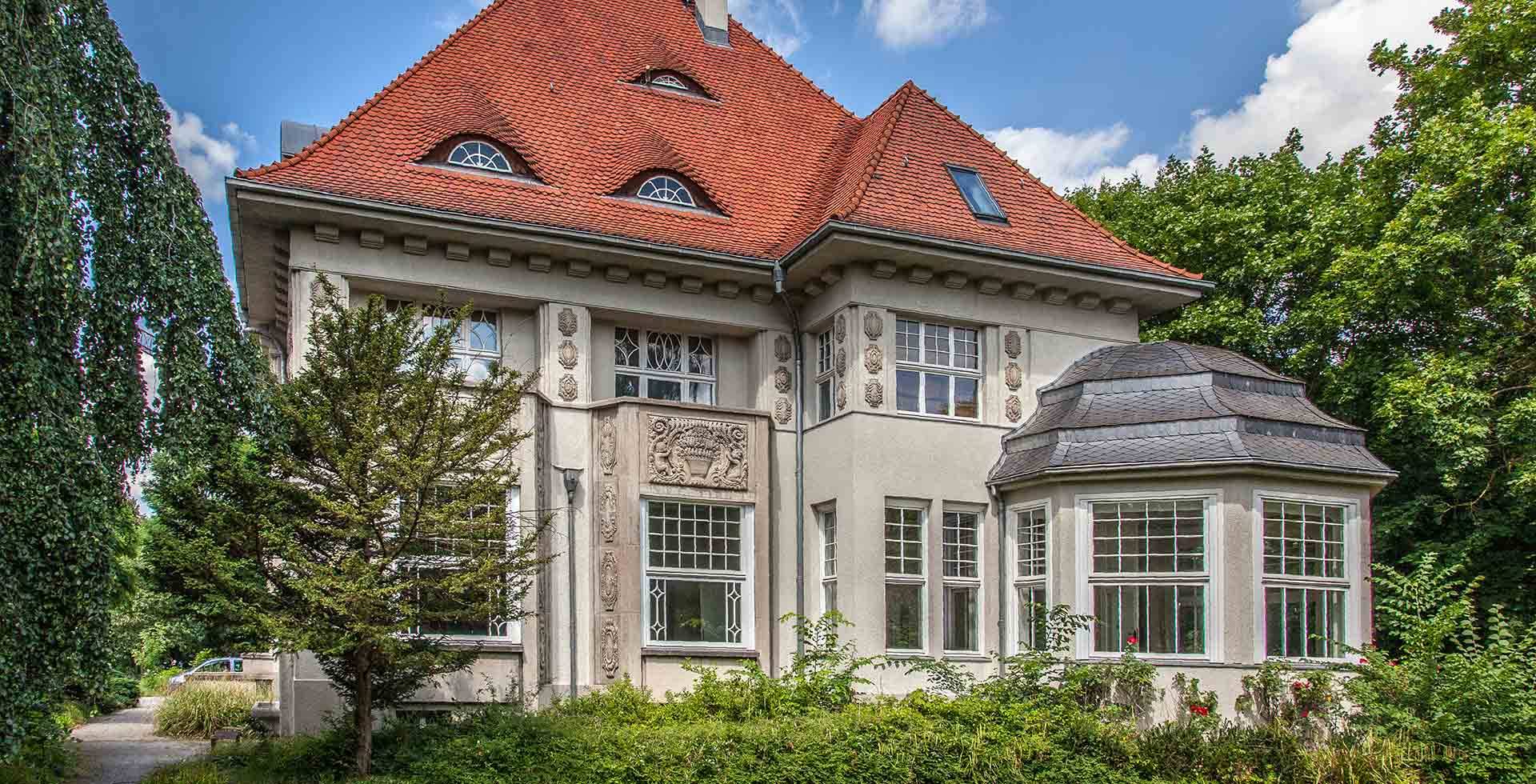 Historische Villa mit roten Ziegeldach, großen Fenstern und dekorativen Steinreliefs an der Fassade.