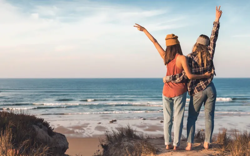 Two friends stand arm in arm and look at the sea, peaceful beach landscape in the background.