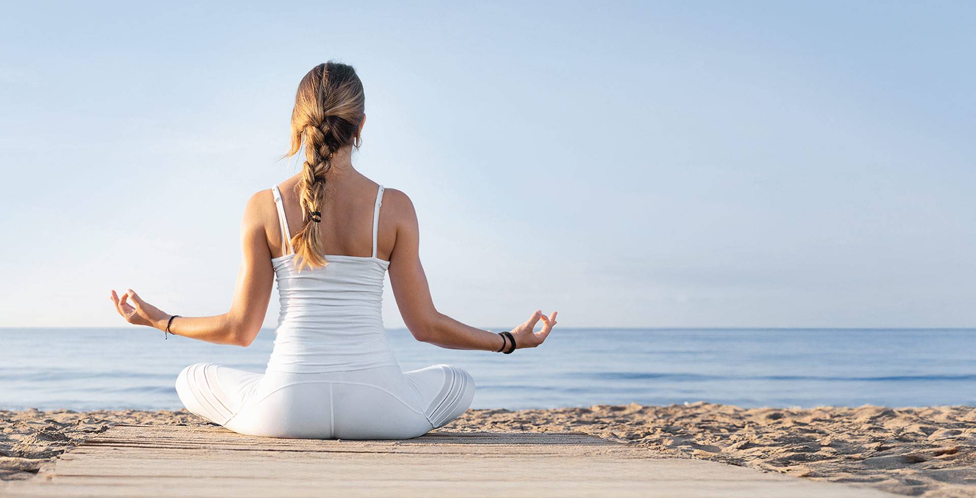 Frau im Schneidersitz beginnt im Sonnenaufgang ihre Yoga-Session am Strand mit Blick aufs Meer.