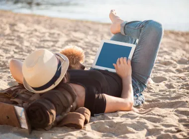 Eine Frau entspannt mit ihrem Pudel am Strand, während sie den arcona Newsletter am Tablet liest.