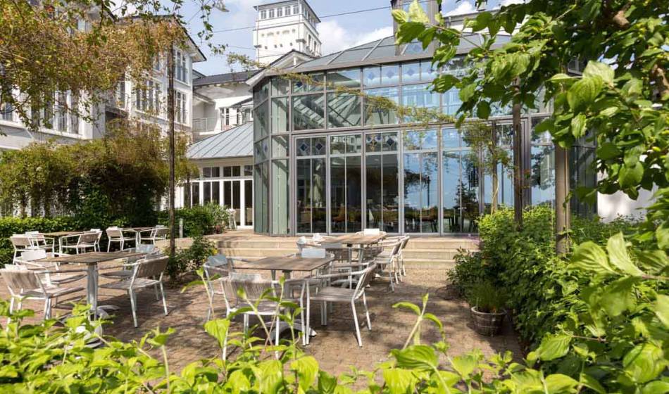 Außenansicht des Strandläufer Restaurants auf Rügen mit moderner Glasfassade, umgeben von Pflanzen und einer möblierten Terrasse.