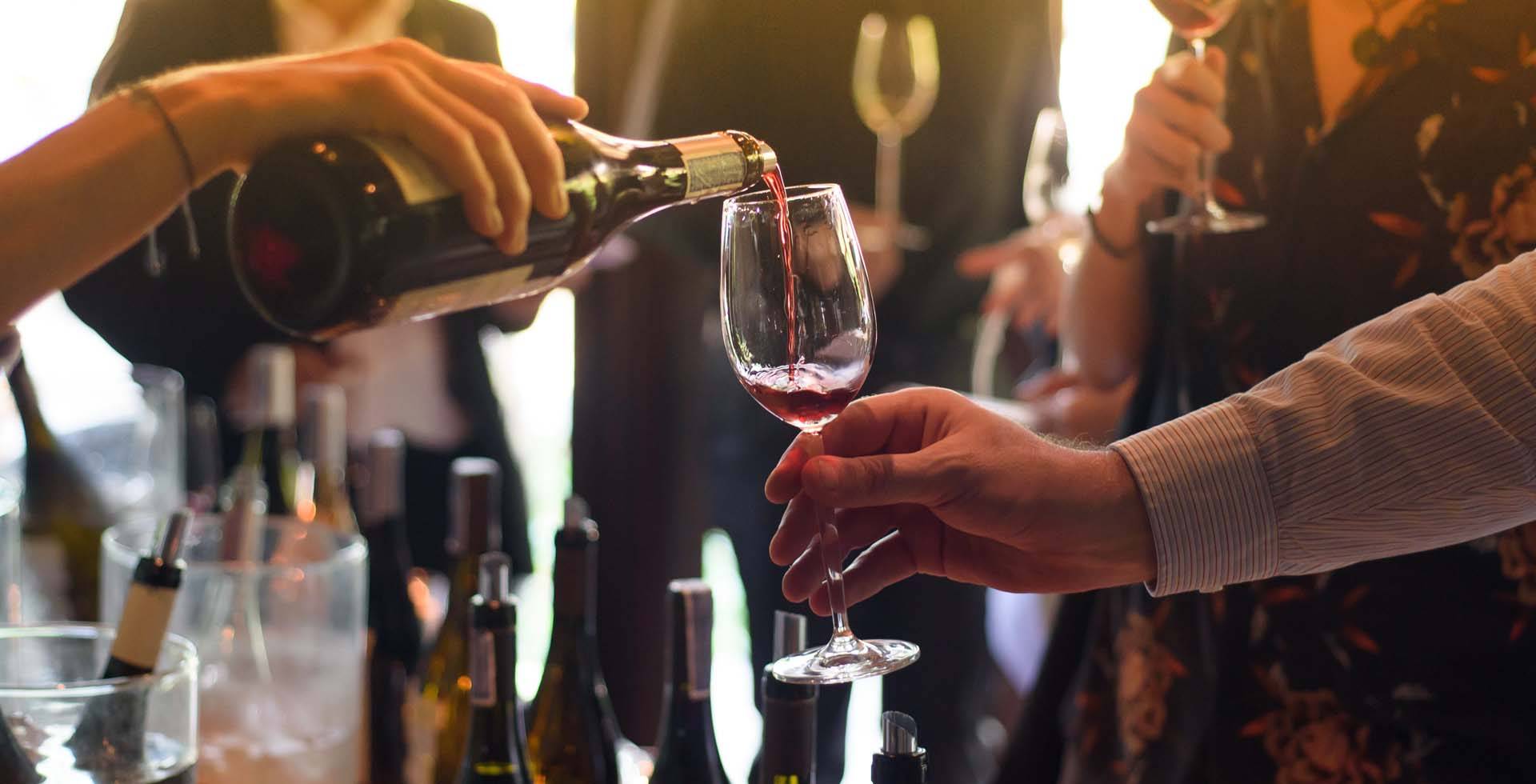 Person schenkt Rotwein aus einer Flasche in ein Glas, weitere Flaschen und Personen im Hintergrund.