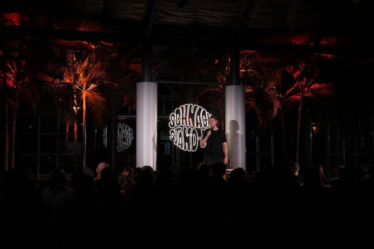 Stand-up-Comedian auf Bühne vor Publikum, Neon-Schriftzug „Schnack Stand-Up“ und Palmen im stimmungsvoll beleuchteten Eventraum im Gasthaus Zur Historischen Mühle Potsdam.