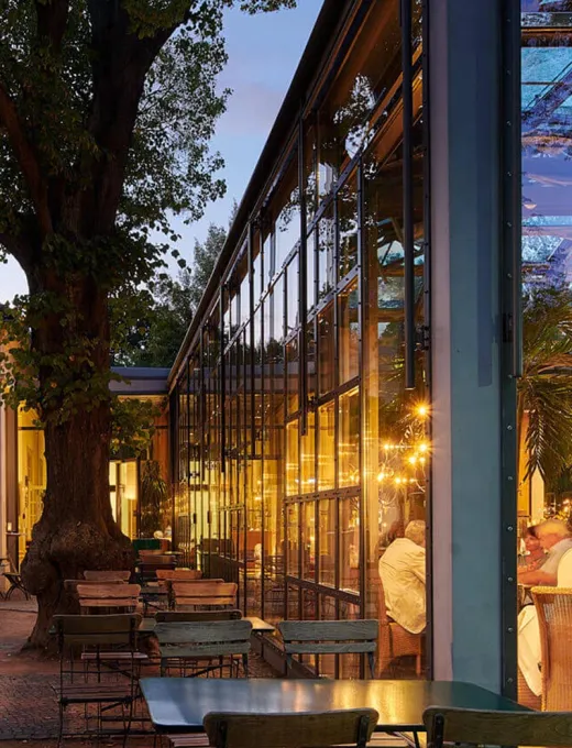 Glass facade of a restaurant at dusk, patio furniture and atmospheric lighting create an inviting atmosphere.