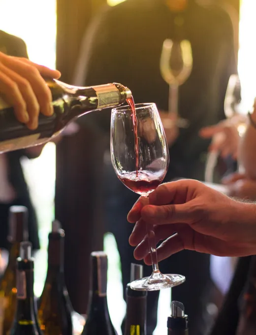Person pouring red wine into a glass at a wine tasting, bottles and guests in the background.