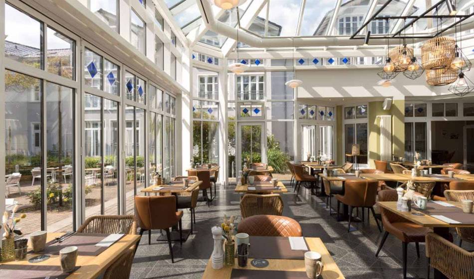 Lichtdurchfluteter Wintergarten im Vju Hotel Rügen, eingerichtet mit Tischen und Stühlen zum Frühstücken.