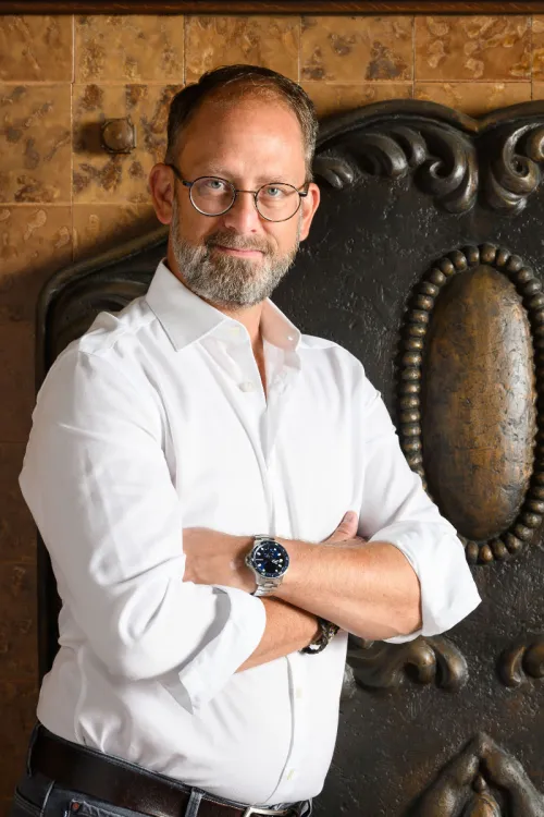 Alexander Winter with glasses and a white shirt stands with his arms crossed in front of a decorative background.