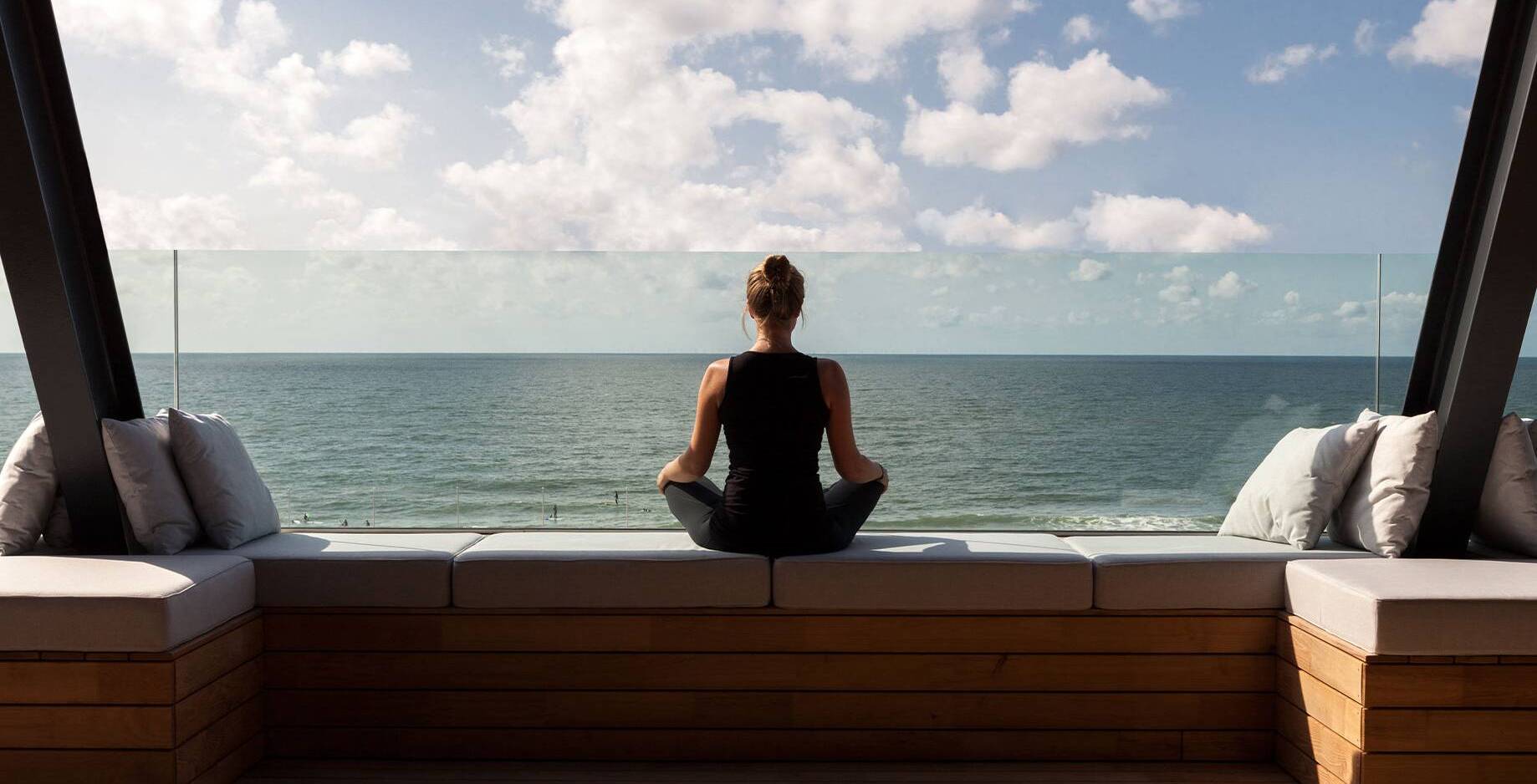 Frau in Yoga-Pose sitzt auf einer Terrasse mit Blick auf das Meer.