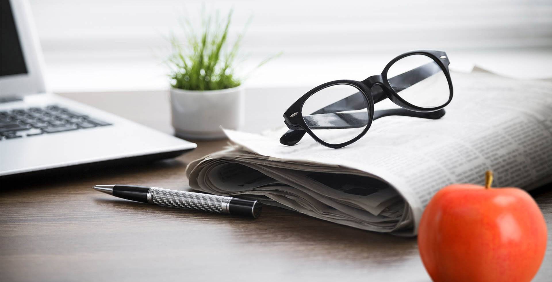 Aufgerollte Zeitung mit Brille, Stift, Laptop und rotem Apfel auf einem Schreibtisch vor einem Fenster.