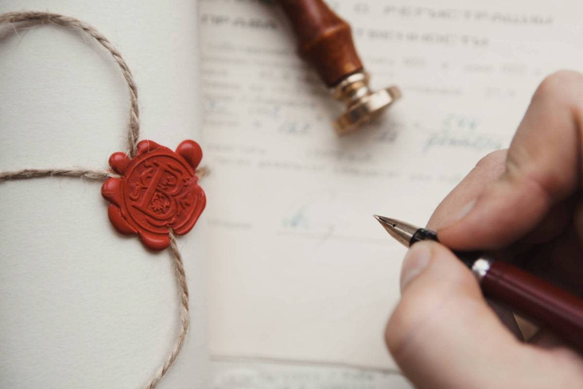 Nahaufnahme eines handgeschriebenen Dokuments mit rotem Wachssiegel und Siegelstempel im Hintergrund. Eine Hand hält einen Füllfederhalter.