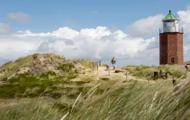 A lighthouse on Sylt, surrounded by dunes and a person with a dog on a walk.