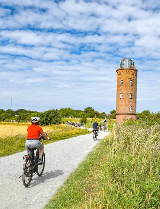 Cyclists on a path in the middle of nature, the lighthouse stands in the background under a blue sky.