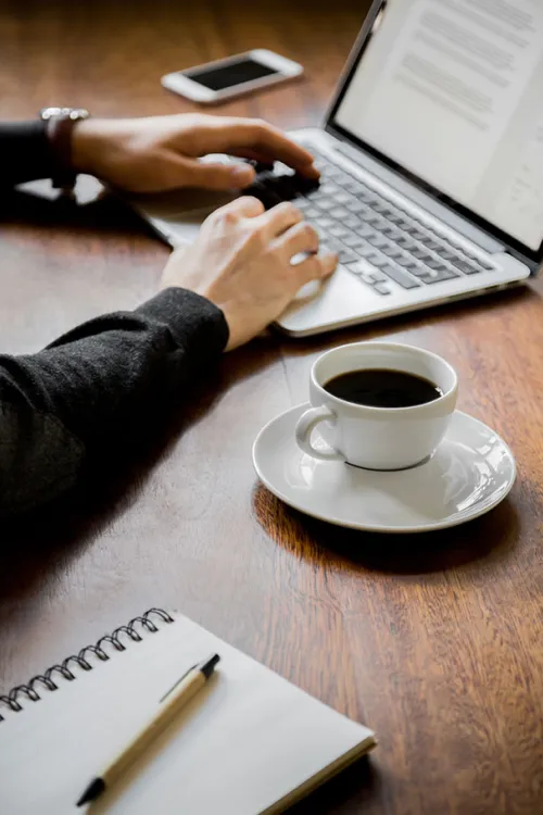 Person typing on laptop, next to a cup of coffee, notepad with pen and smartphone on a wooden table in the Arcona Hotels & Resorts.