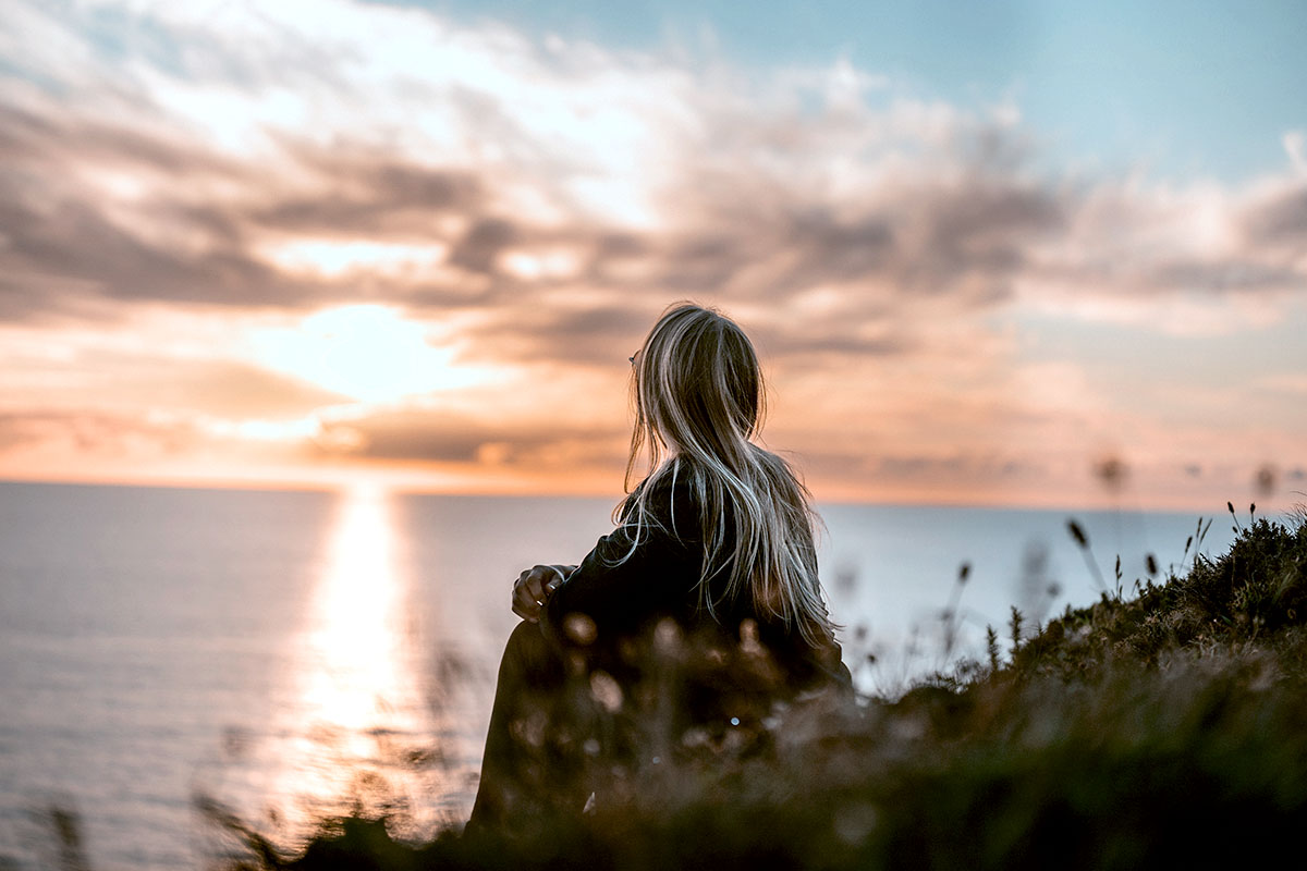 Eine junge Frau mit blondem Haar sitzt an einer Stranddüne auf Rügen und genießt den farbenfrohen Sonnenuntergang im Ostseeurlaub.