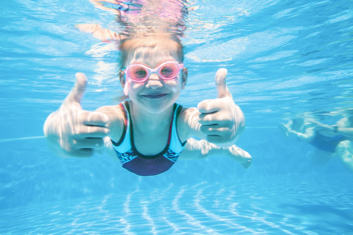 Fröhliches Kind mit Schwimmbrille taucht im Pool und zeigt beide Daumen hoch beim Schwimmkurs im Vju Hotel auf Rügen.