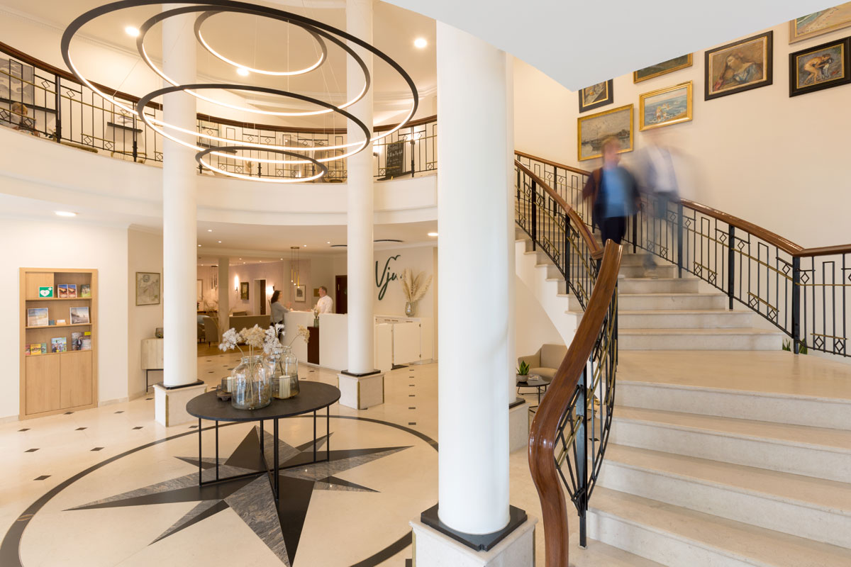 Einladender Empfangsbereich mit heller Lobby, geschwungener Treppe und stilvollen Kronleuchtern im Vju Hotel auf Rügen.