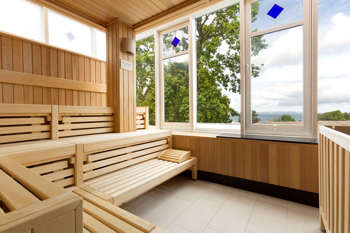 Helle Holzsauna mit Bänken, großen Fenstern und Blick auf die Ostsee und grüne Natur im Wellnessbereich des Vju Hotels.