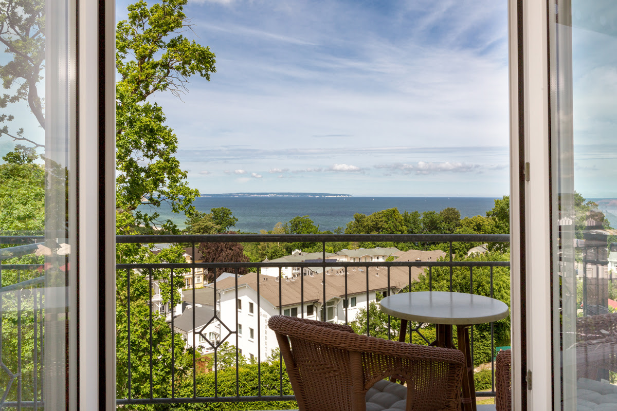 Geöffnete Balkontür mit Aussicht auf die Ostsee, Bäume und Häuser im Sonnenlicht vor dem Vju Hotel auf Rügen.
