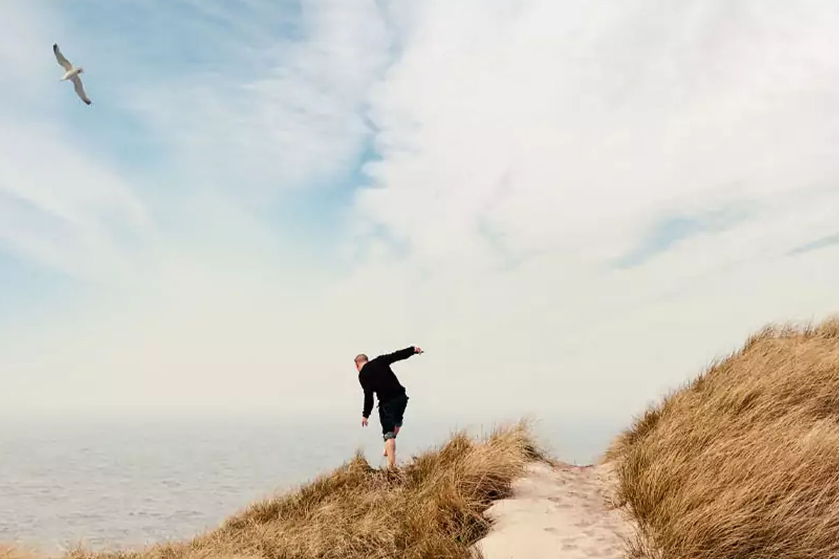 Ein Mann balanciert auf einem Dünenweg bei windigem Herbstwetter mit Blick auf das Meer im Herbsturlaub im Wyn. Strandhotel.