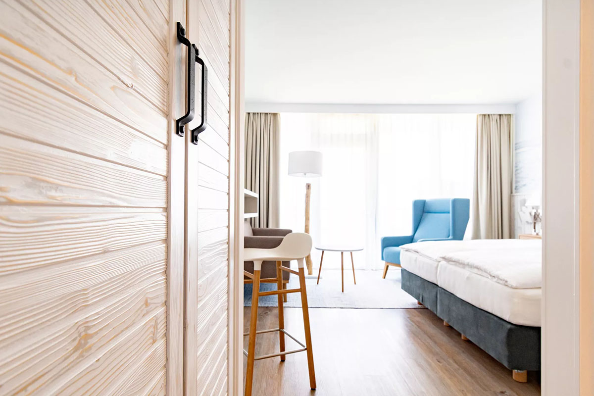 Einladendes Zimmer Meerzeit im Wyn. Strandhotel mit blauem Sessel und gemütlichem Bett, lichtdurchflutet durch bodentiefe Fenster.