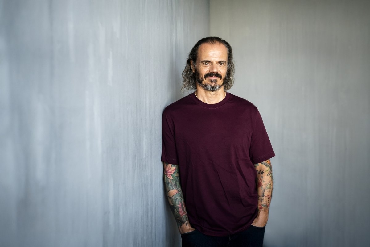 Mann mit schulterlangen Haaren und Bart, trägt ein bordeauxfarbenes T-Shirt, steht entspannt mit verschränkten Armen vor einer grauen Wand, tätowierte Unterarme sichtbar.