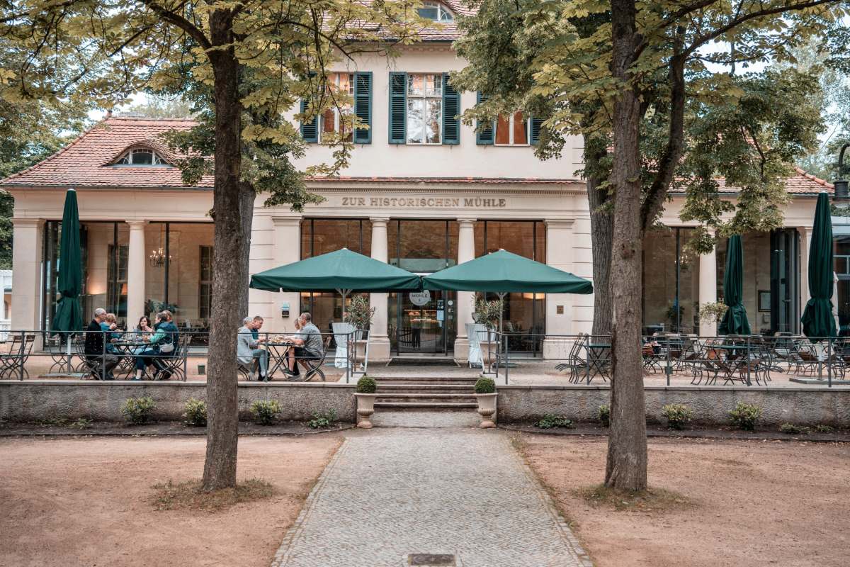 Aussenansicht des Restaurants Zur historischen Mühle in Potsdam mit großer Terrasse, grünen Schirmen und Gästen.