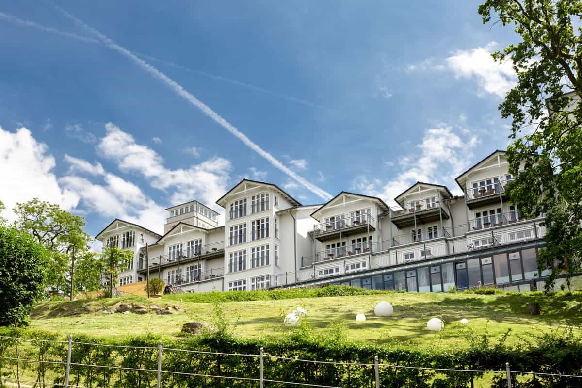 Außenansicht des Vju Hotels mit klassischer Architektur, großen Fenstern und Balkonen, gelegen auf einer grünen Anhöhe mit gepflegter Landschaft.