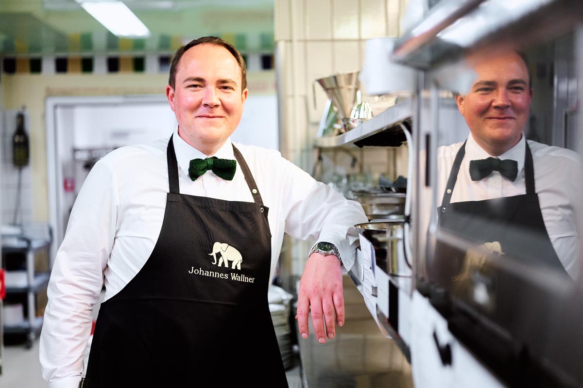Chef Johannes Wallner stands smiling in a professional kitchen and leans relaxed against a work surface.