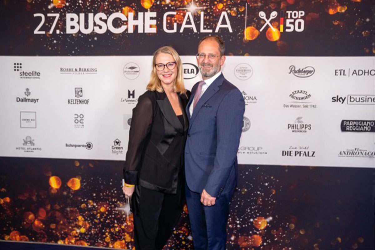 Eine Frau und ein Mann stehen lächelnd vor einer Wand und und freuen sich über eine Auszeichnung.