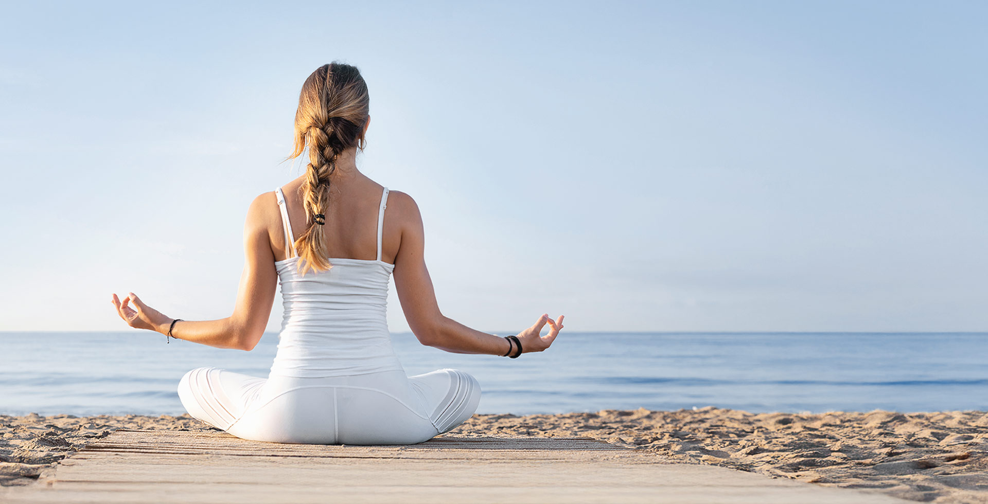 Frau im Schneidersitz beginnt im Sonnenaufgang ihre Yoga-Session am Strand mit Blick aufs Meer.