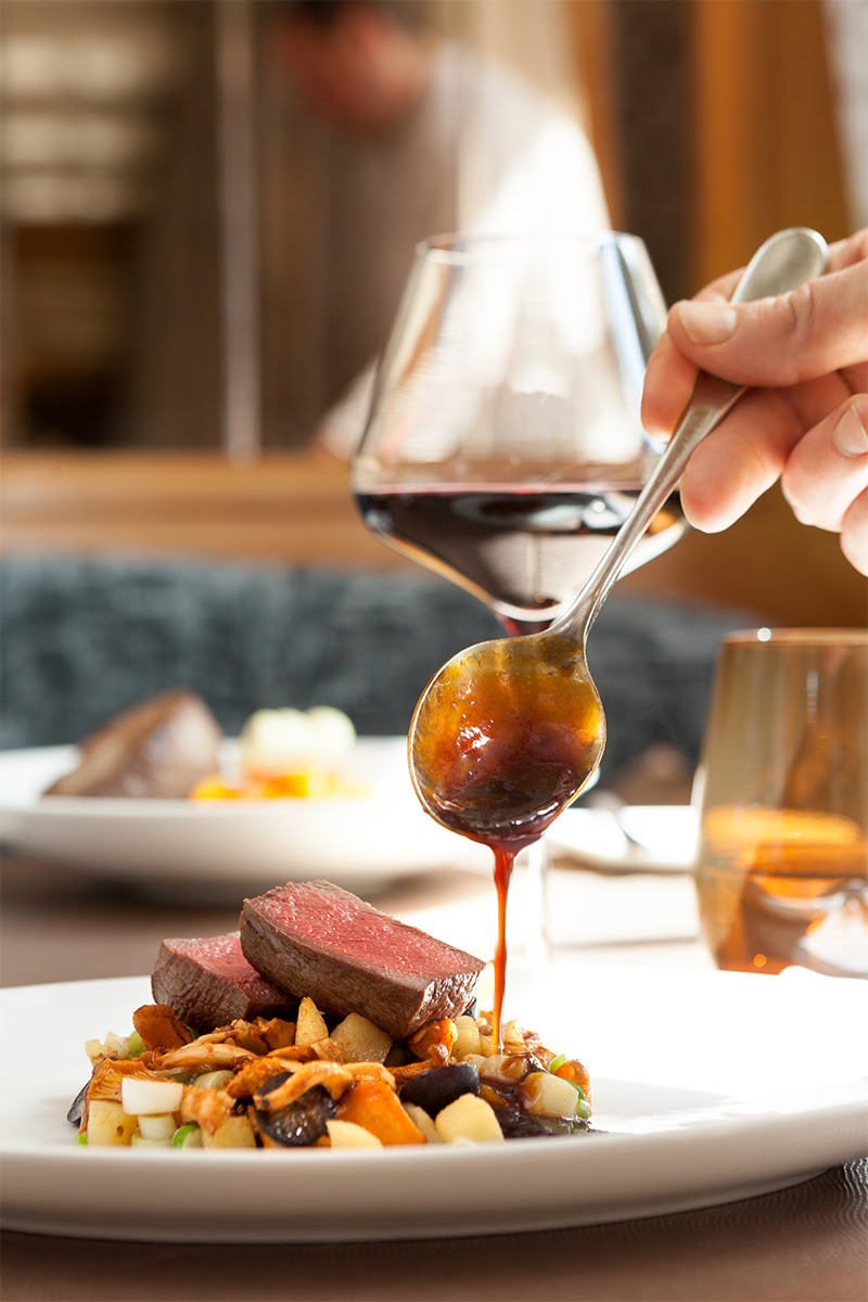 Hand eines Kochs, der eine Sauce über ein Stück Fleisch auf einem Teller mit Gemüse gibt, dazu ein Glas Rotwein auf dem Tisch.