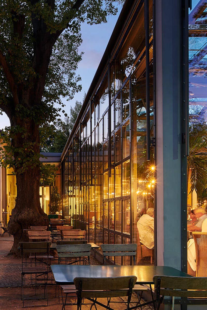 Glasfassade eines Restaurants bei Abenddämmerung, Terrassenmöbel und stimmungsvolle Beleuchtung schaffen eine einladende Atmosphäre.