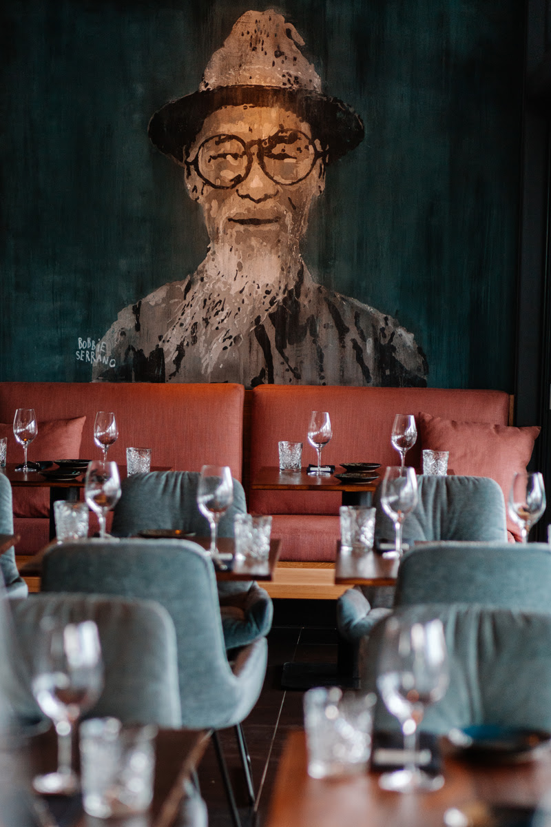 Restaurant mit gedeckten Tischen, Weingläsern und einer großen Wandmalerei eines Mannes mit Hut und Brille im Hintergrund.