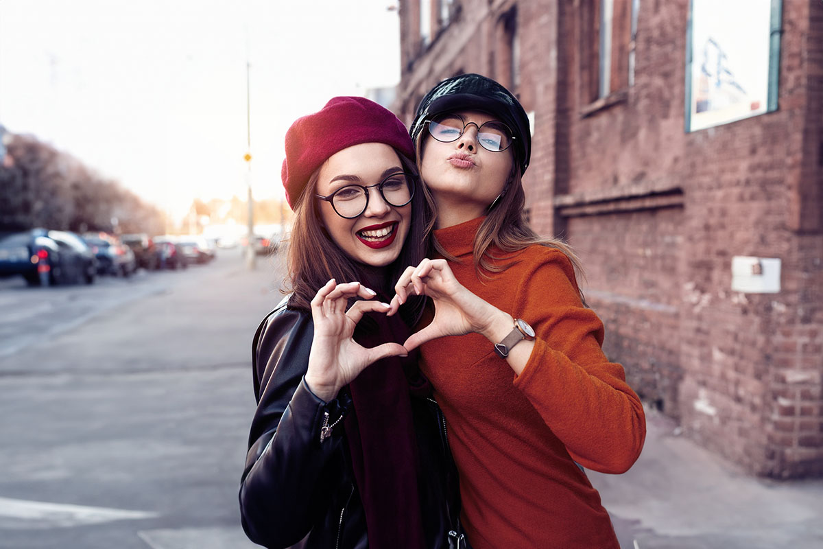 Freundinnen lächeln in die Kamera, Herzgeste symbolisiert Freundschaft und Zusammenhalt.