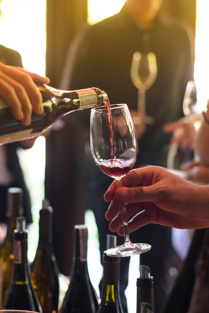 Person schenkt Rotwein in ein Glas bei einer Weinverkostung ein, Flaschen und Gäste im Hintergrund.