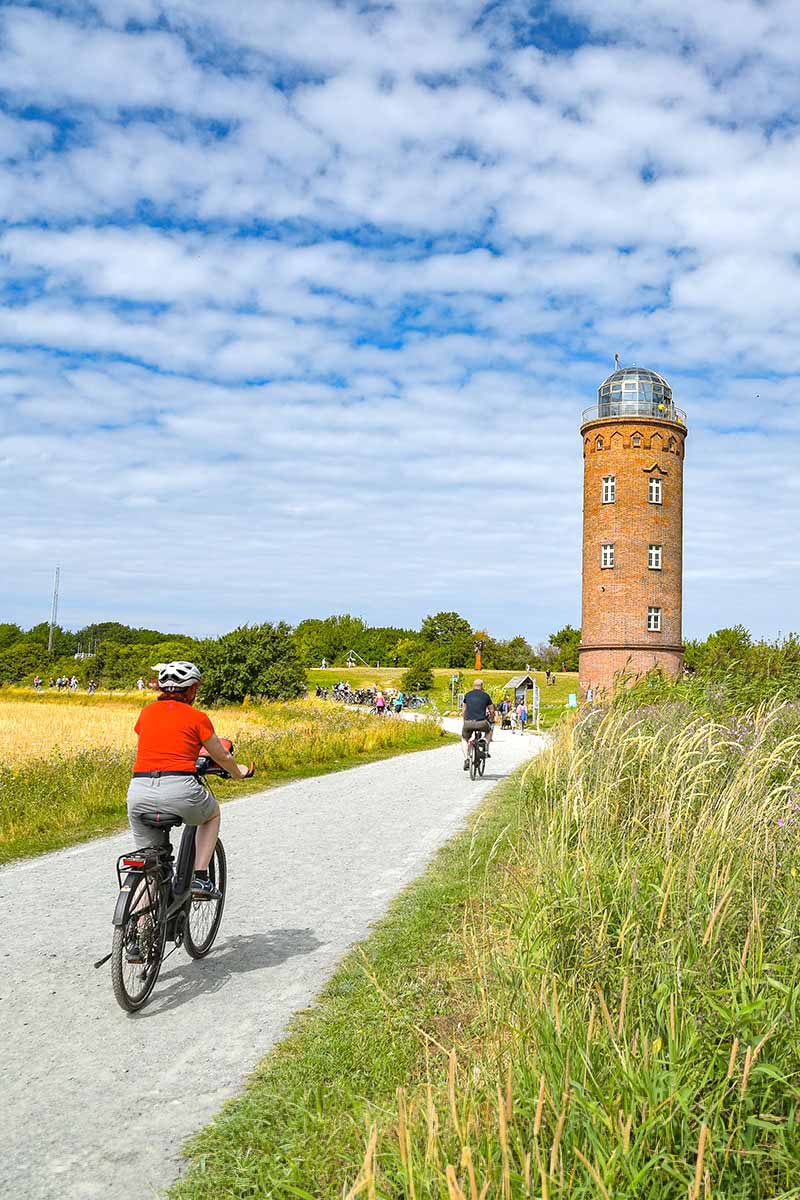 Fahrradfahrer auf einem Weg inmitten der Natur, der Leuchtturm steht im Hintergrund unter blauem Himmel.