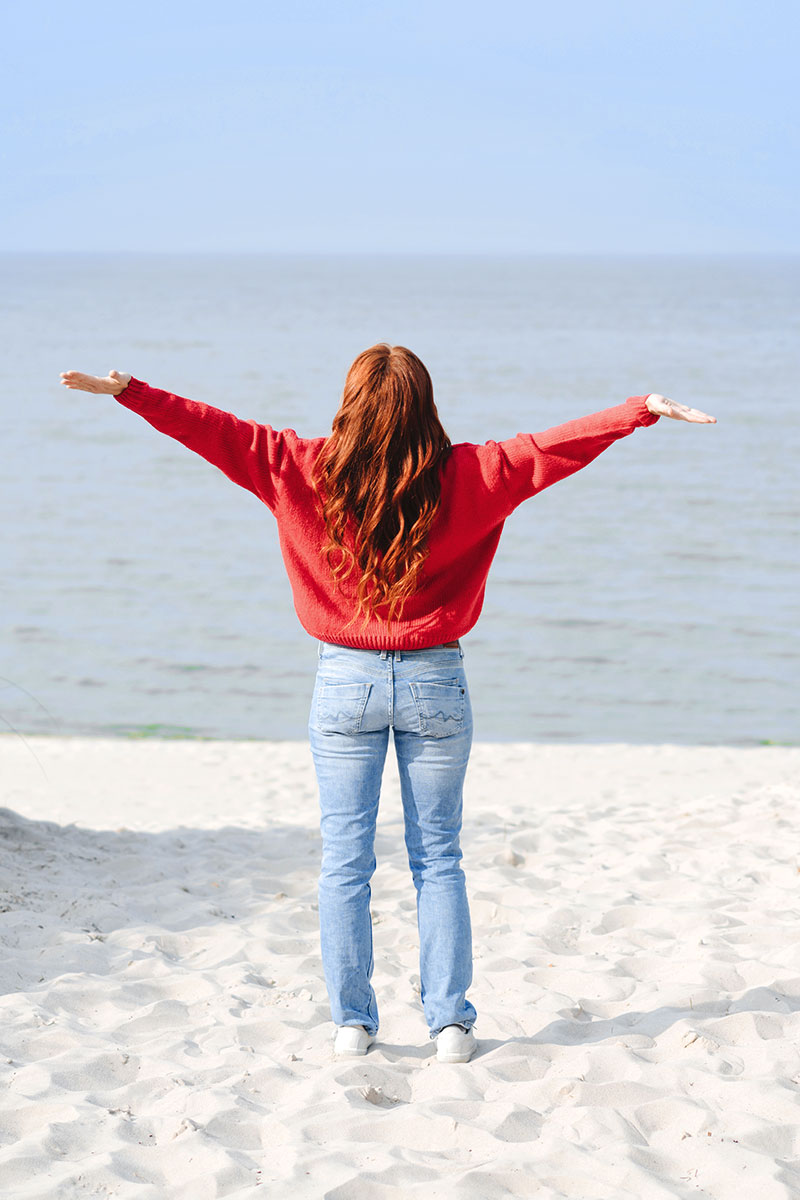 Frau in rotem Pullover steht mit ausgestreckten Armen am Strand und blickt aufs Meer.