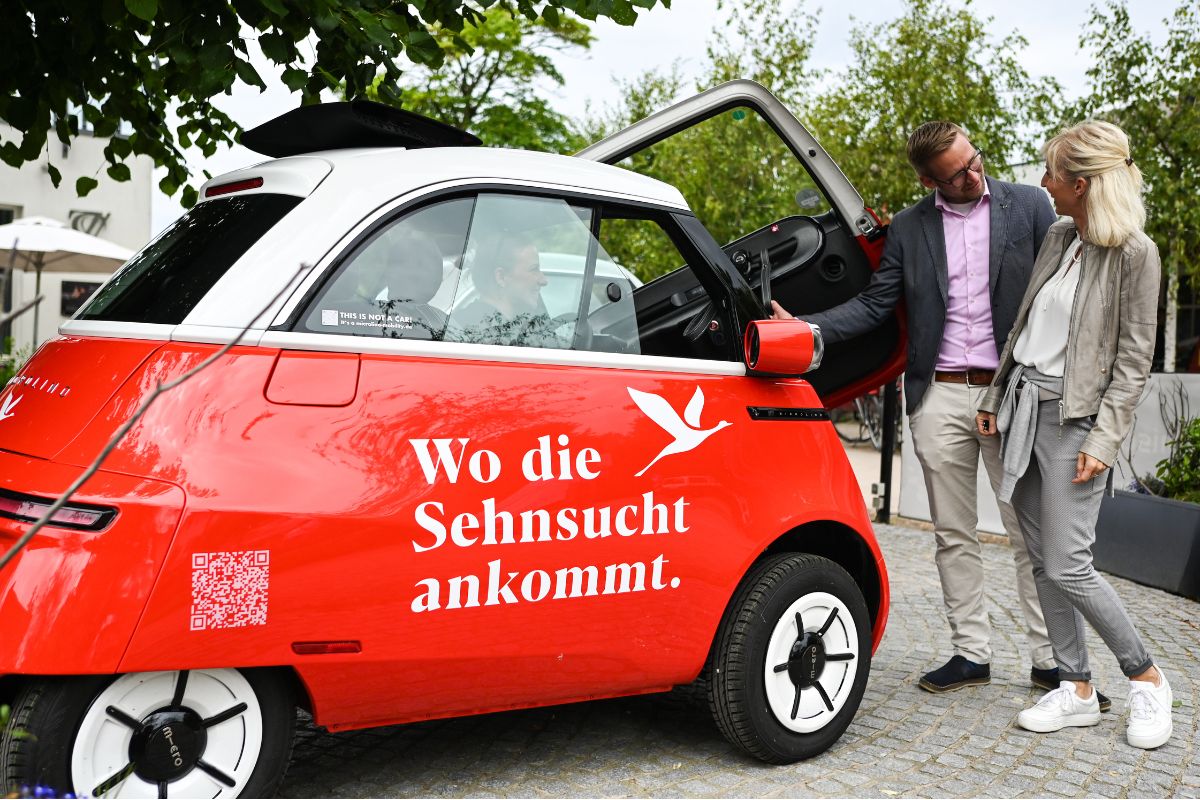 Zwei Personen stehen neben einem roten Microlino mit der Aufschrift Wo die Sehnsucht ankommt.