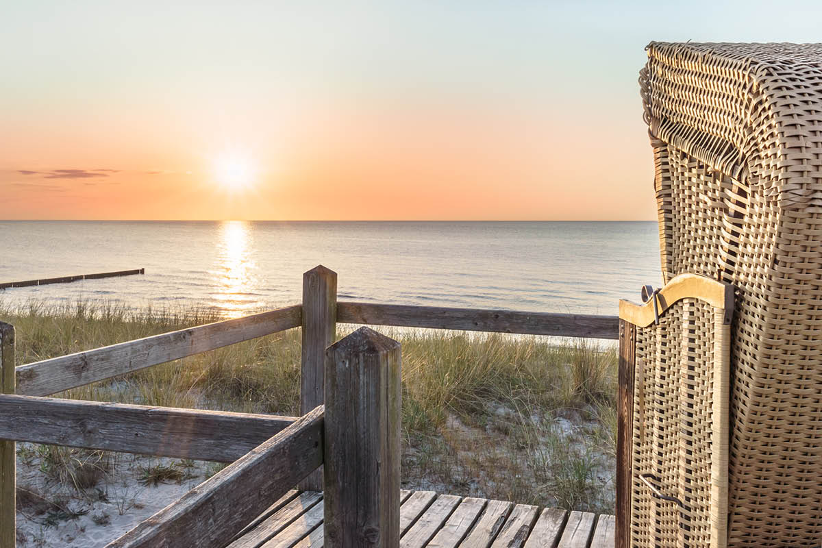 Sonnenuntergang an der Ostsee, Strandkorb auf Holzsteg, umgeben von Dünen, während die Sonne das Meer golden faerbt