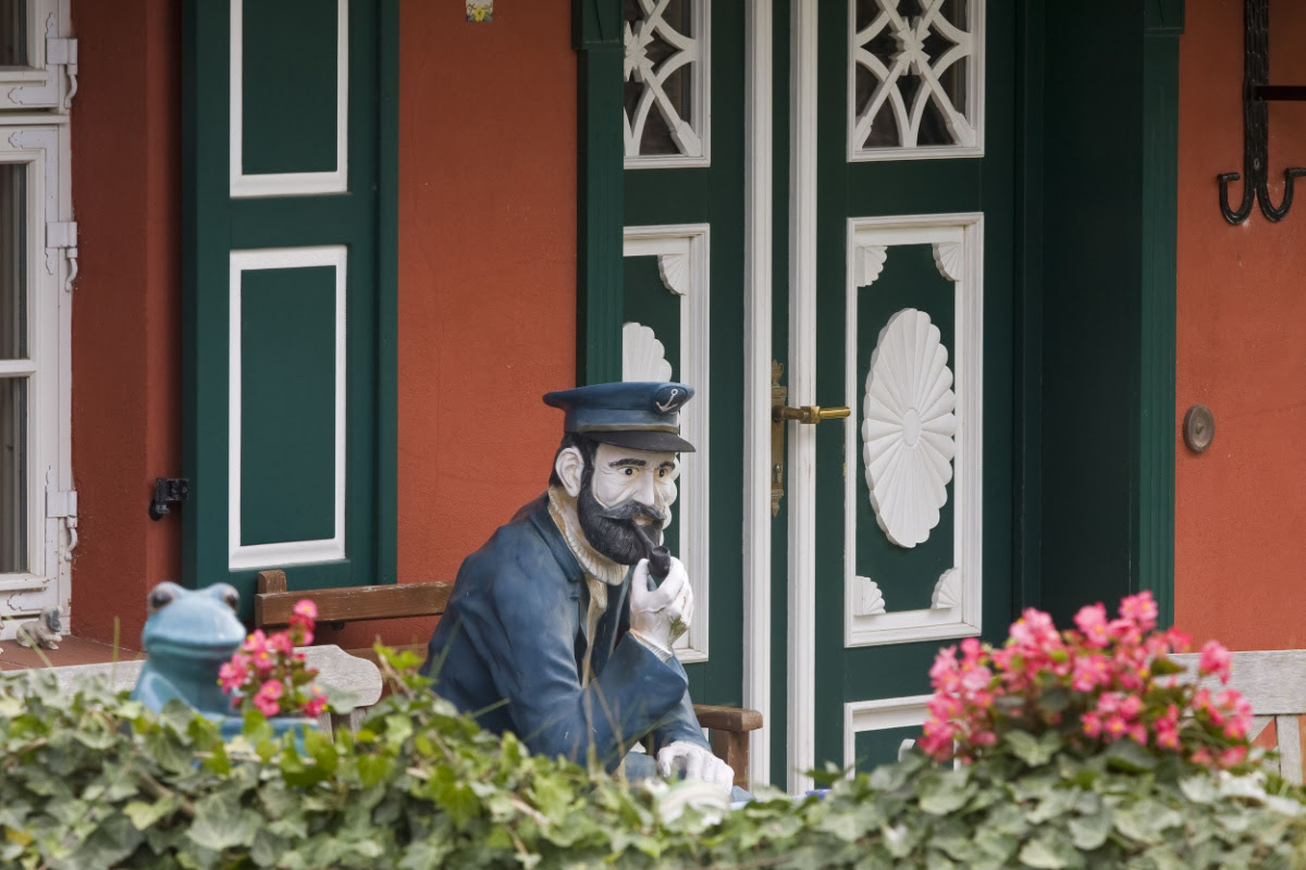 Statue eines Seemanns mit Bart und Mütze vor einem rot-grünen, verzierten Haus, umgeben von Blumen und Efeu