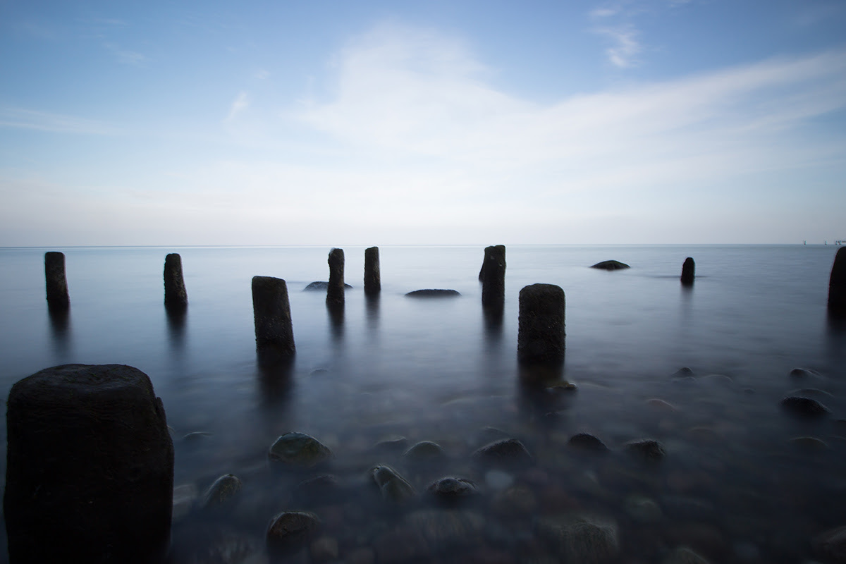 Mehrere Holzpfeiler stehen im klaren Wasser der ruhigen Küstenlandschaft auf Rügen, unter blauem Himmel.